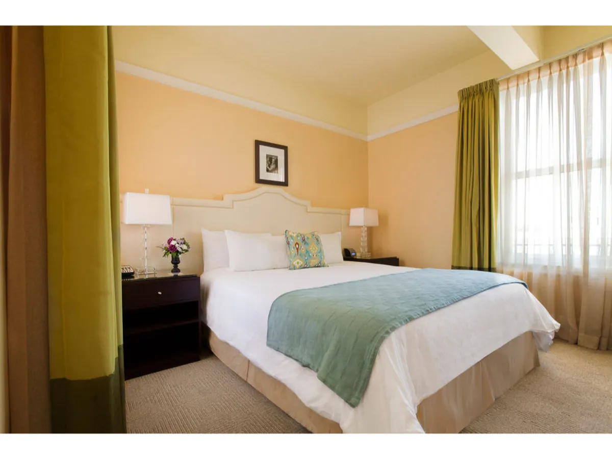 The image showcases a beautifully styled hotel room with a plush bed adorned with decorative pillows, bathed in natural light streaming through a large window. The inviting setup creates a peaceful ambiance, perfect for relaxation or enjoying a leisurely morning by the window.