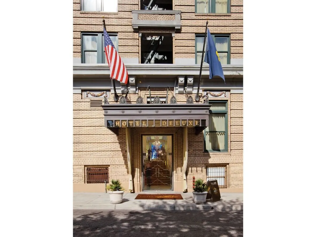 A welcoming exterior shot of the hotel’s entrance, featuring the Hotel Deluxe sign, a set of international flags, and the grand doorway. The inviting approach is framed by tasteful architectural details and inviting lighting, emphasizing luxury and a sense of arrival.