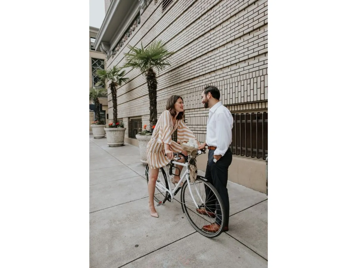An exterior scene of a couple preparing for a bike ride, with a bicycle and vibrant potted plants against a charming brick wall. The image captures a playful, romantic moment and nods to the property’s complimentary bike rentals for urban exploration.