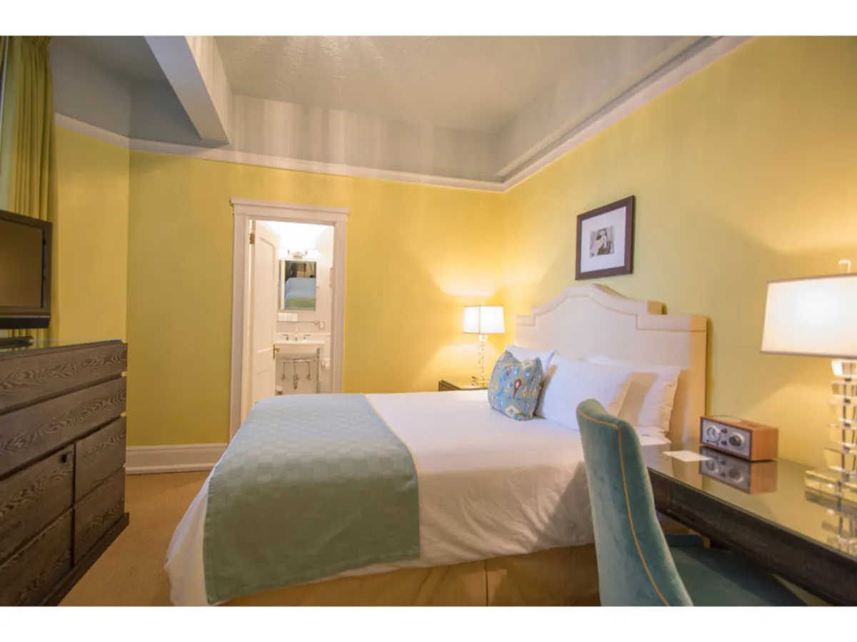 A calming bedroom scene featuring a plush bed with decorative pillows, a bedside lamp casting a warm glow, a dressing table with a mirror, and a dresser topped with a television. Sunlight filters through the window, illuminating the restful space that blends classic elegance with modern comforts.