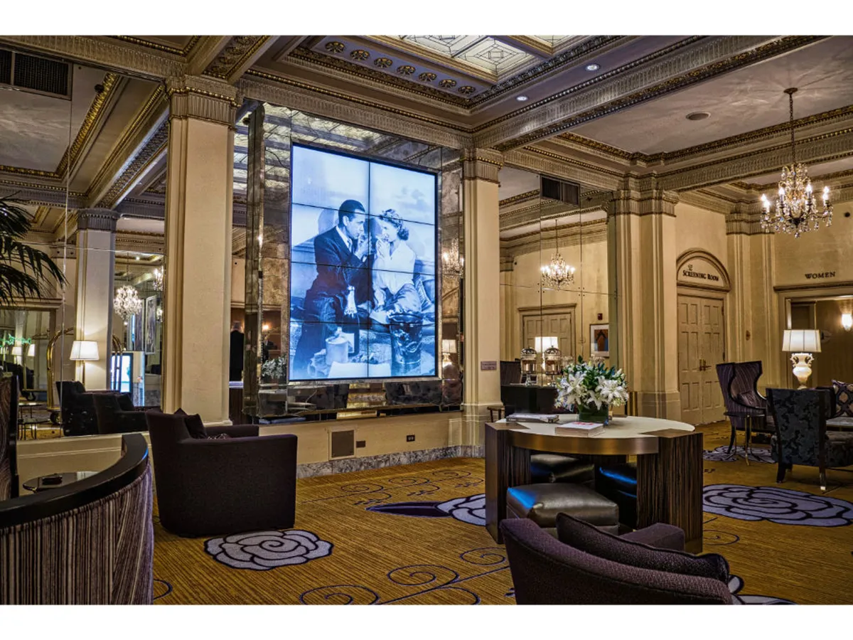 A luxe, ornately decorated interior with a vintage chandelier, intricately detailed ceiling, and a prominent digital display showing a classic black-and-white photograph. Stylish, comfortable seating invites conversation and relaxation in an atmosphere that seamlessly blends glamour and nostalgia.