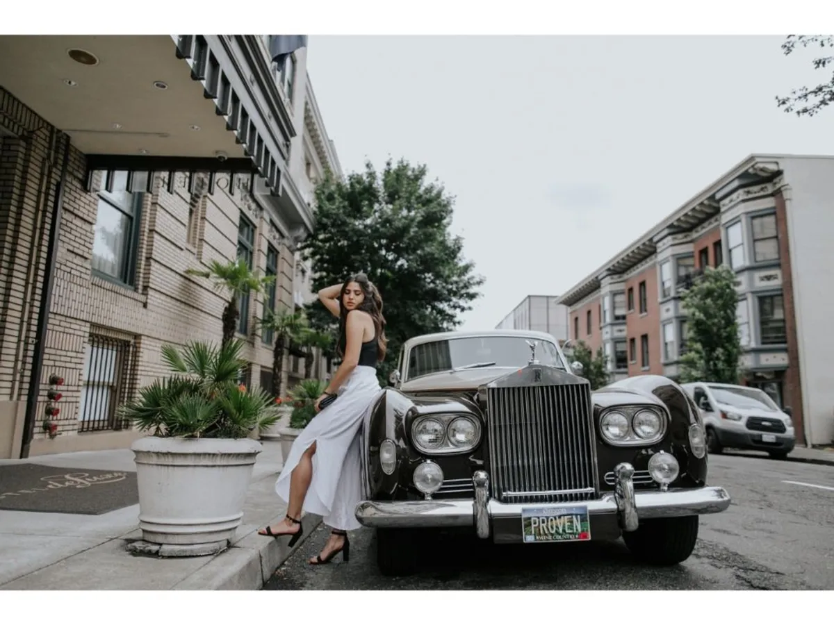 An exterior scene featuring a classic car parked along an urban street, with a young woman in stylish attire nearby. The backdrop includes elegant urban architecture and lush planters, evoking a sense of city exploration and fashionable leisure. The image captures the sophisticated ambiance and walkable charm highlighted in the section.