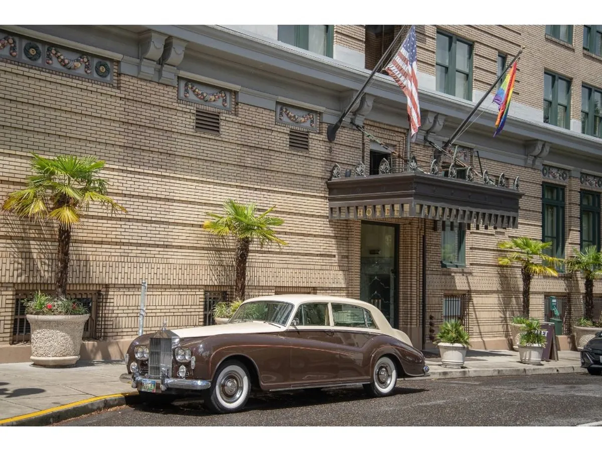 This image shows the grand exterior entrance of Hotel Deluxe, featuring an ornate awning, palm trees, waving flags, and a classic Rolls Royce parked out front. The historic building’s facade is illuminated, conveying a sense of sophisticated arrival and luxury hospitality in a vibrant downtown setting.