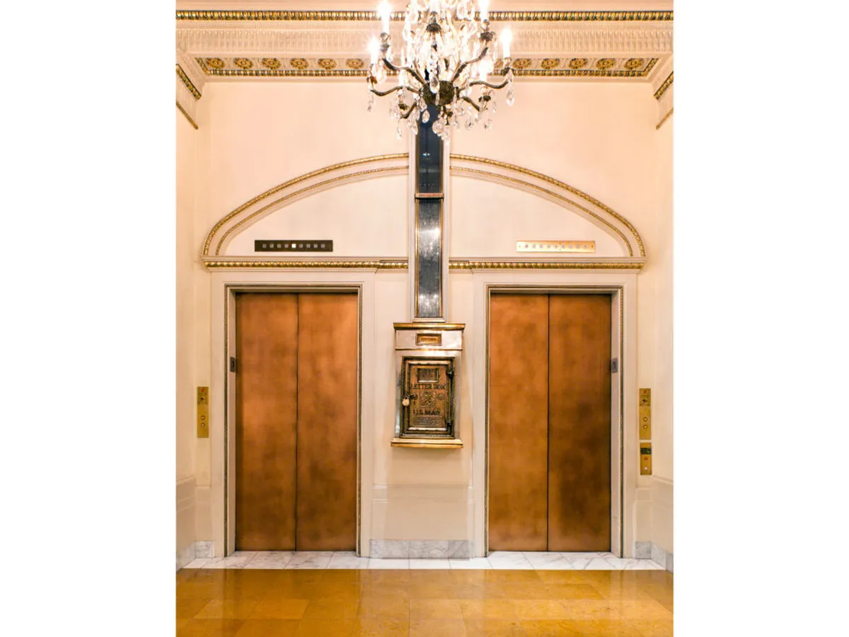 A sophisticated hotel lobby with ornate chandeliers, decorative wall details, and elevator doors. The space exudes a timeless elegance and offers a welcoming area for guests, including those traveling with pets.