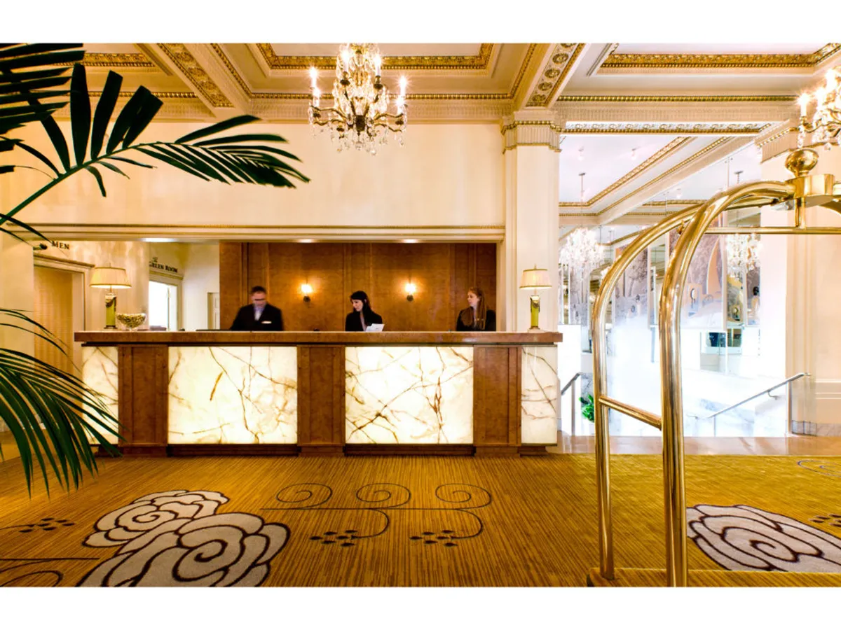 The image features a sophisticated hotel reception area. Staff members stand behind an elegant front desk under a sparkling chandelier, while a decorative plant adds a touch of warmth. The setting exudes professionalism and hospitality, indicating attentive service and a welcoming environment.