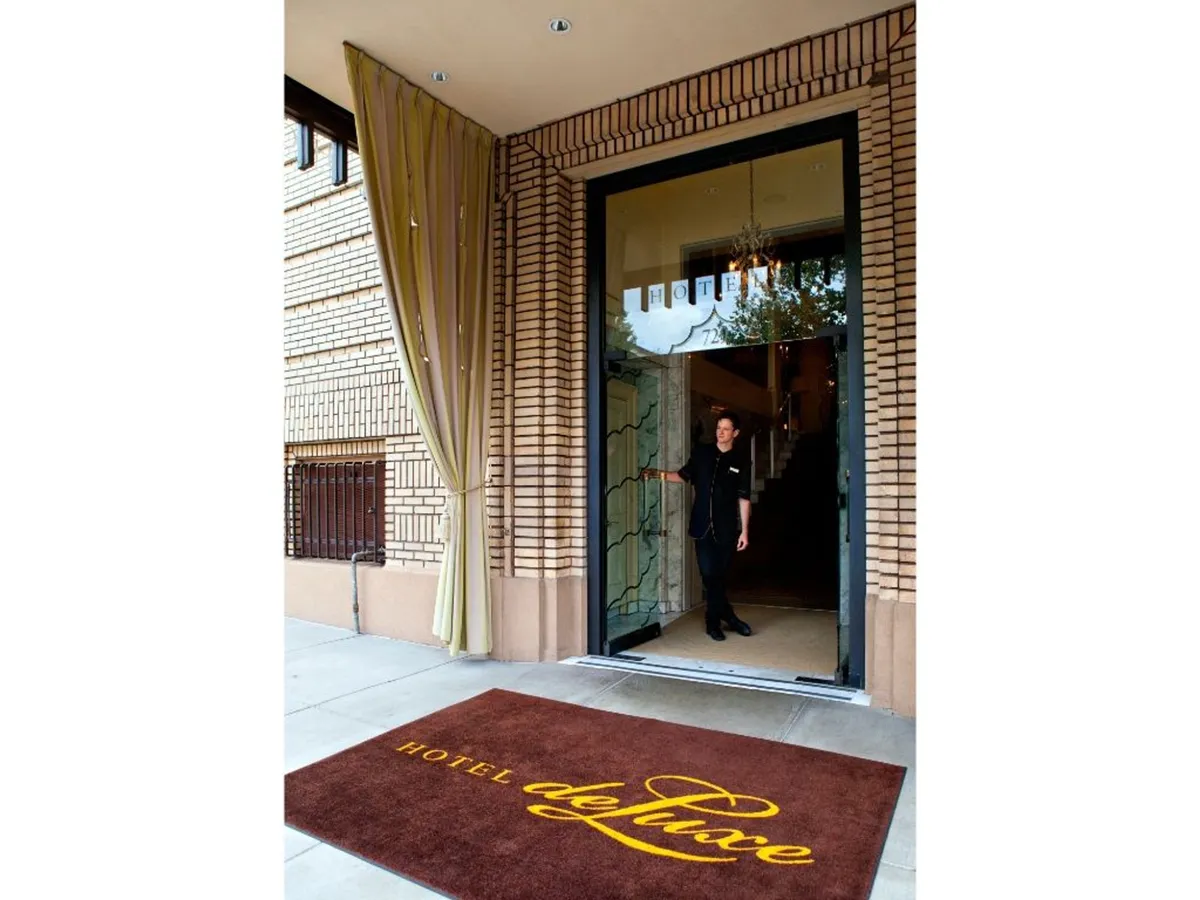 This image highlights the welcoming entrance to Hotel Deluxe, featuring the entrance door, a branded welcome mat, and hotel staff ready to greet arriving guests. The scene is warm and personal, suggesting attentive service and a seamless check-in experience.