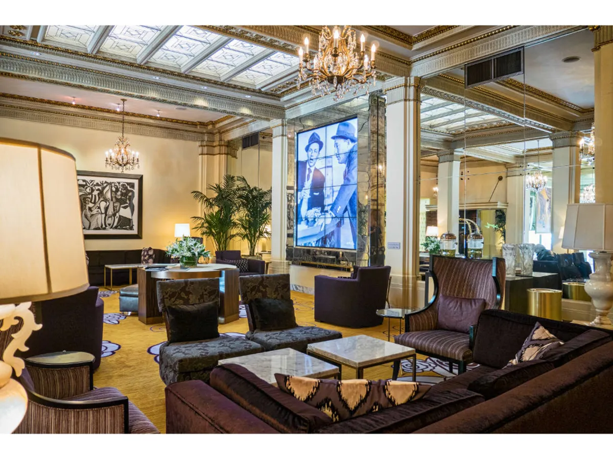 A sophisticated hotel interior featuring a sparkling chandelier, plush seating arranged in an inviting lounge area, and a large screen displaying historical film art. Decorative plants add a touch of nature, and the atmosphere suggests a space for both socializing and quiet art appreciation.
