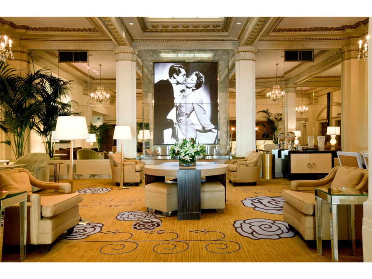 A beautifully arranged cocktail lounge featuring a large black and white artwork of a couple, a prominent central floral arrangement, and plush, luxurious seating designed for intimate conversations and gatherings. The space exudes Old Hollywood glamour with artistic touches.
